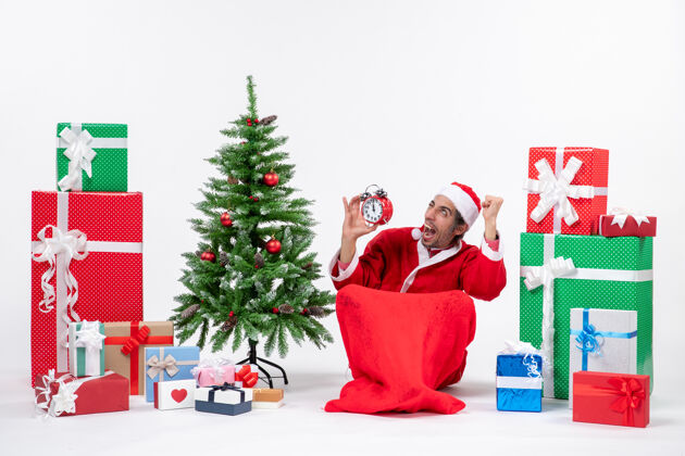 圣诞树情绪激动 高兴 惊讶 疯狂的圣诞老人坐在地上 在礼物和装饰圣诞树旁的白色背景上展示钟表地面坐着惊讶