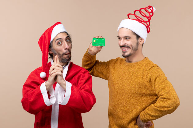 两个正面图两个快乐的家伙 一个穿着圣诞老人外套 正在做手指枪 另一个手放在米色孤立背景的腰部两个快乐的家伙信贷外套