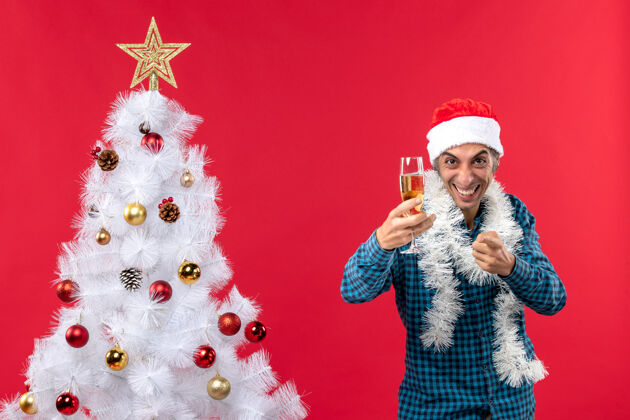 圣诞老人带着圣诞老人帽 穿着蓝色条纹衬衫 拿着一杯葡萄酒 指着圣诞树旁边的东西的年轻人的圣诞心情什么的圣诞人