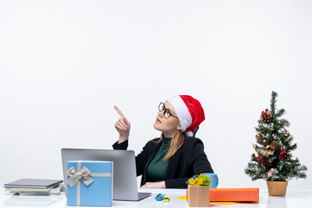 秘书一个戴着圣诞老人帽子和眼镜的严肃的年轻女人坐在一张桌子旁 桌子上有一棵圣诞树和一份白色背景的礼物笔记本电脑帽子圣诞老人