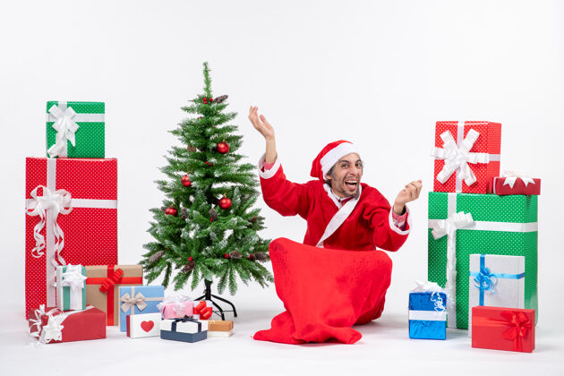 右边惊讶有趣的年轻人打扮成圣诞老人与礼物和装饰圣诞树坐在地上指着白色背景的右边的东西地上礼物装饰