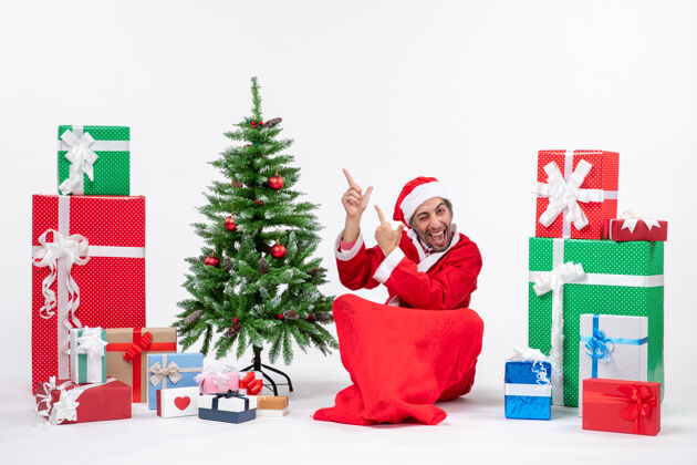 圣诞老人有趣的年轻人打扮成圣诞老人与礼物和装饰圣诞树坐在地上指着白色背景的右边的东西礼物坐着成人