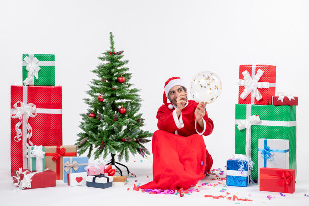 年轻圣诞气氛与年轻有趣的圣诞老人坐在圣诞树和礼物在白色背景上不同的颜色圣诞老人微笑圣诞老人