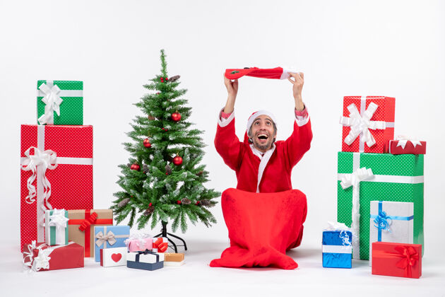 礼物微笑着的圣诞老人坐在地上 把圣诞袜举到头上 放在礼物旁边 在白色背景上装饰新年树提高微笑礼物