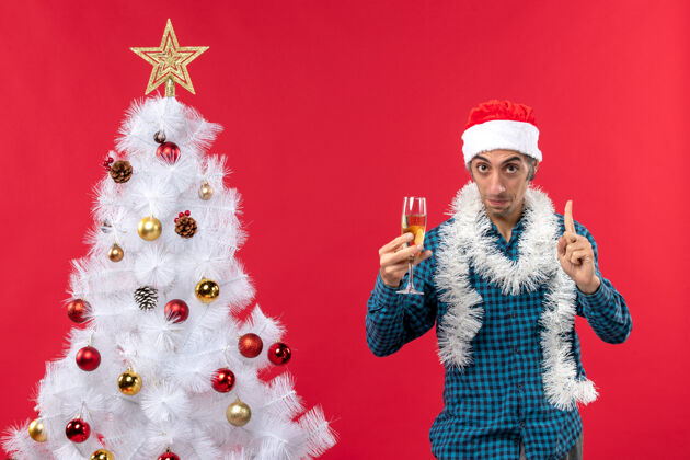 前夜圣诞心情坚定的年轻人戴着圣诞老人的帽子 穿着蓝色条纹衬衫 在圣诞树旁举起一杯葡萄酒葡萄酒年轻人玻璃