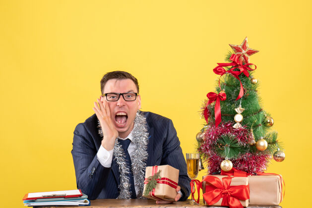 男人一个年轻人对着坐在圣诞树旁的桌子上的人大喊大叫 并把礼物穿上黄色坐着办公室树
