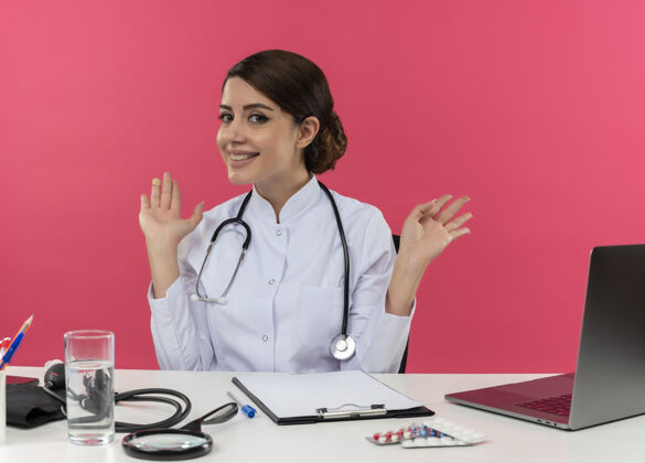 女高兴的年轻女医生穿着医用长袍 手持听诊器坐在办公桌旁 用医疗工具在电脑上工作 用复印空间摊开双手医生电脑长袍