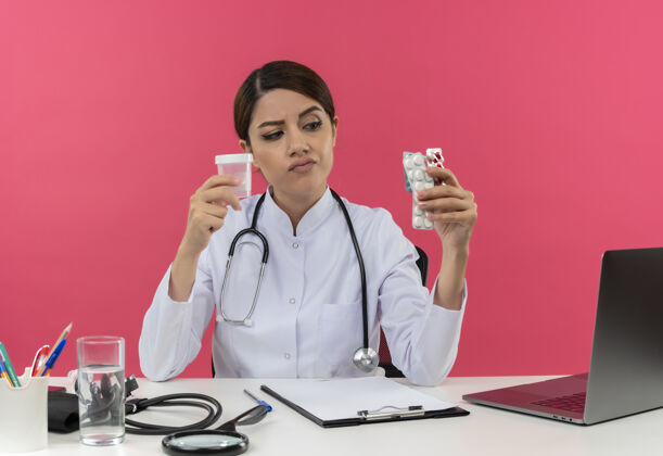 药片想着年轻的女医生穿着医用长袍 带听诊器 坐在办公桌旁 拿着医疗工具 拿着空罐子和药片 还有复印空间空听诊器工具