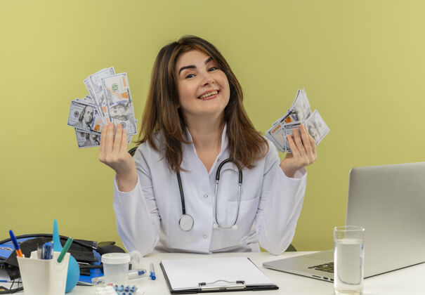 长袍微笑的中年女医生穿着带听诊器的医用长袍坐在办公桌旁用笔记本电脑和医疗工具拿着现金和复印空间医疗工具笔记本电脑