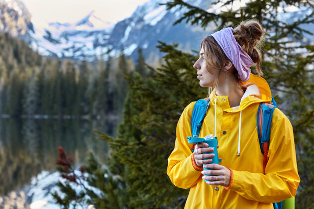 杉木沉思的女登山者头戴围巾 穿黄色雨衣 从烧瓶里喝热咖啡放松水远足
