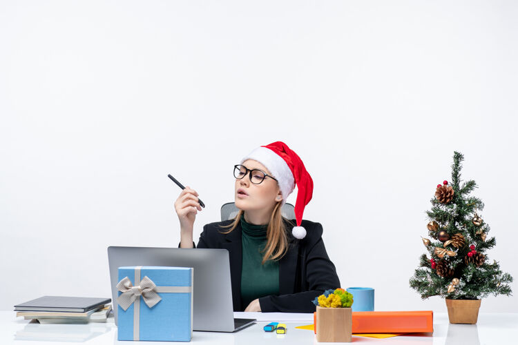 金发女郎新年的气氛与集中金发女人与圣诞老人的帽子坐在一张桌子上 圣诞树和一个礼物在它的白色背景金发女郎女人秘书