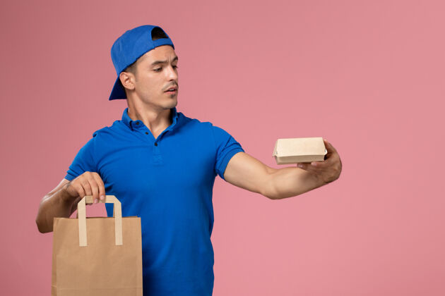 送货正面图：身穿蓝色制服和披风的年轻男性快递员 手上拿着快递包裹 站在粉红色的墙上前面人包裹