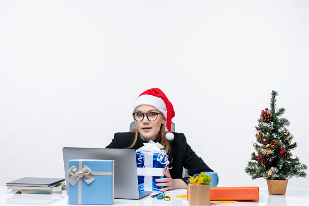 笔记本电脑带着圣诞心情的年轻女子戴着圣诞老人的帽子 戴着眼镜坐在一张桌子旁 把礼物和圣诞树装饰在它的白色背景上坐着年轻人礼物