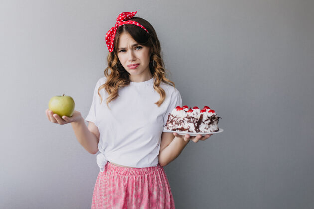苹果苗条悲伤的女孩拿着水果和蛋糕迷人的卷发女模特不能决定吃什么苗条波浪人