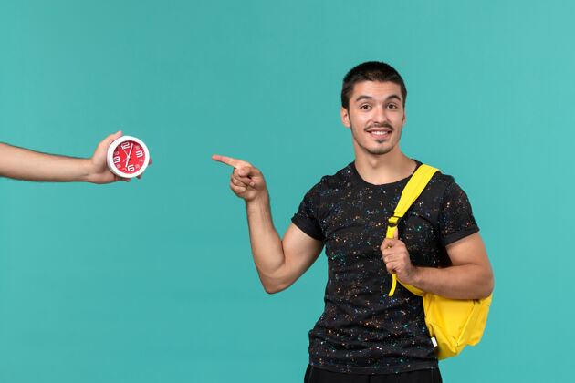 男身穿深色t恤黄色背包的男学生在浅蓝色墙上微笑的正面视图大学微笑游戏设备