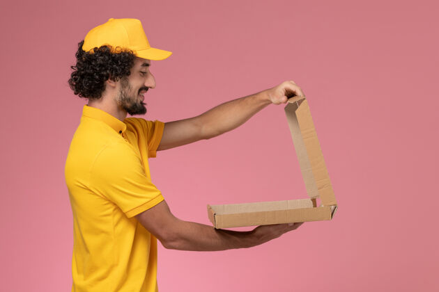 食物正面图：身穿黄色制服的男快递员拿着食物盒站在浅粉色的墙上男快递盒子前面
