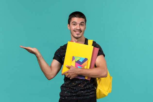 书前视图中的男学生穿着深色t恤黄色背包 在浅蓝色的墙上拿着文件和抄写本男男学生大学
