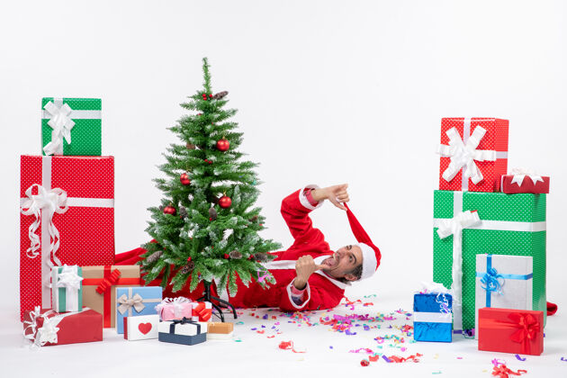 说谎在白色背景下 年轻疯狂的圣诞老人躺在圣诞树后的礼物旁 庆祝节日的气氛圣诞礼物年轻