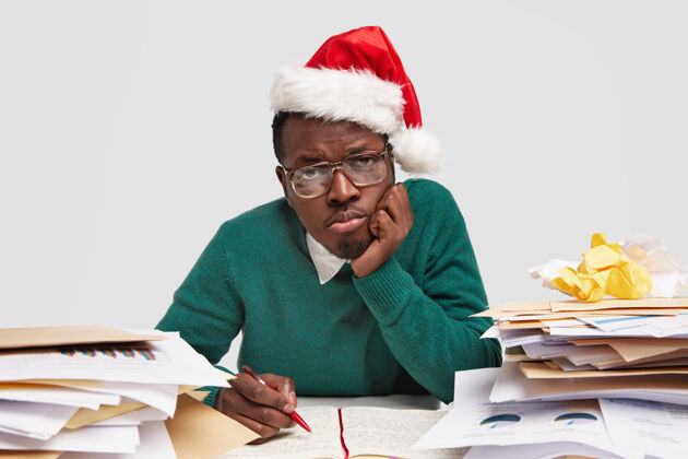 工人过度劳累悲伤的非洲裔美国人手贴脸颊 戴圣诞老人帽 戴眼镜 写日记压力工作狂桌面