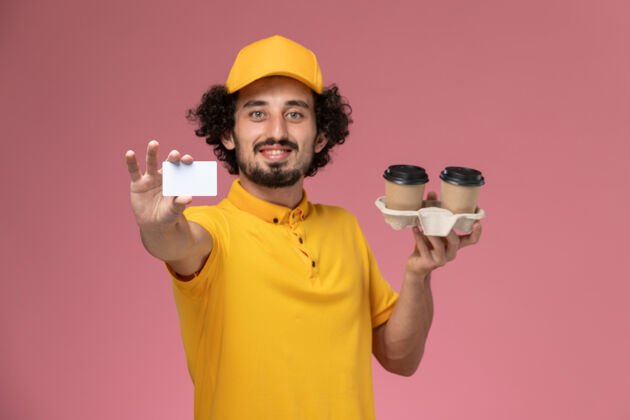 男士正面图：身穿黄色制服和披风的男性快递员手持浅粉色墙上的棕色送货咖啡杯和卡片快递卡片咖啡