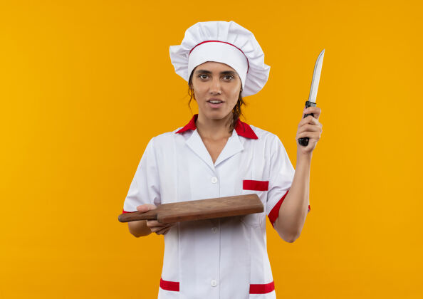 板子年轻的女厨师穿着厨师制服拿着砧板和刀站在隔离的黄色墙壁上 有复印空间拿着制服切