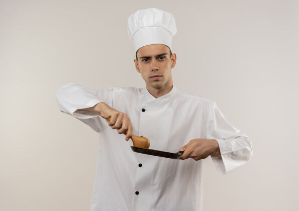 拿着自信的年轻男厨师穿着厨师制服 拿着煎锅和勺子站在孤零零的白墙上男煎着勺子