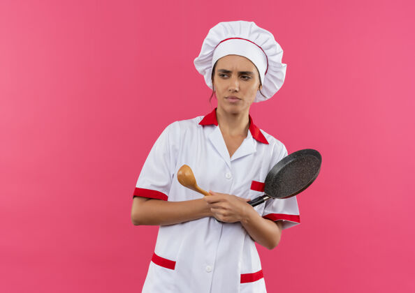 侧看着旁边穿着厨师制服的年轻女厨师 拿着勺子和煎锅 放在粉红色的墙壁上 还有复印空间厨师穿煎