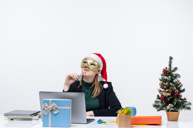 坐着圣诞节的心情 年轻女子戴着圣诞老人的帽子 戴着眼镜 戴着面具 坐在一张桌子旁 在白色的背景上沉思年轻女人圣诞节笔记本