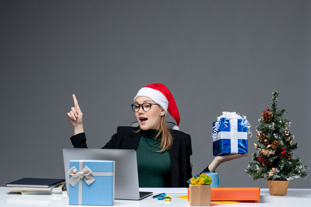 坐着圣诞气氛积极的年轻女子戴着圣诞老人的帽子 戴着眼镜 坐在一张桌子旁 在黑暗的背景上指着上面的礼物笔记本电脑圣诞老人眼镜