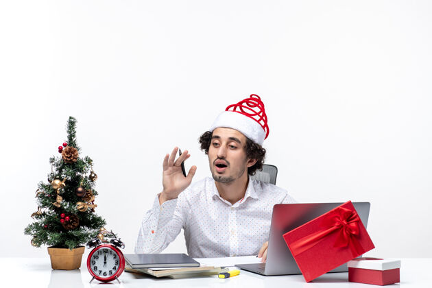 帽子困惑的年轻商人 戴着滑稽的圣诞老人帽 在白色背景的办公室里检查笔记和庆祝圣诞节年轻圣诞老人笔记