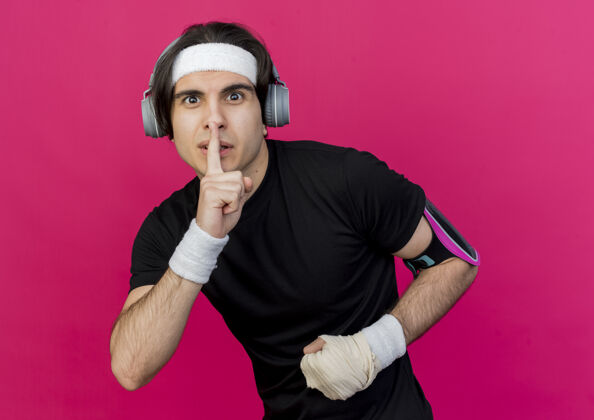 制作年轻的运动型男子 戴着运动服和头带 戴着手机和智能手机袖标 手指放在嘴唇上做着沉默的手势运动装站着年轻