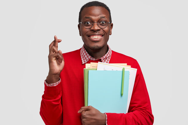 惊讶照片中幸福的黑人男子交叉手指祈求好运 背着课本 有着牙齿般的微笑 穿着红色的衣服非洲毛衣牙齿