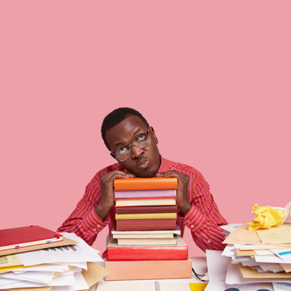 知识不高兴的美国黑人学生的照片 闷闷不乐的表情 手放在一堆教科书上 歪着头 穿着粉色衬衫眼镜家庭作业大学