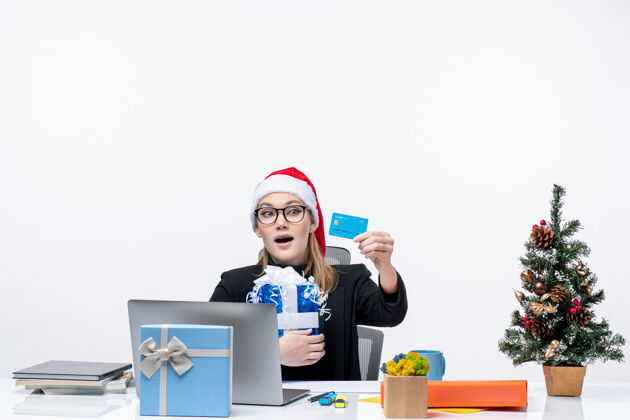 圣诞老人戴着圣诞老人帽 戴着眼镜的快乐年轻女子坐在桌子旁 手里拿着圣诞礼物 看着白色背景上的银行卡帽子眼镜圣诞老人