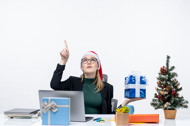 圣诞节带着圣诞老人帽 戴着眼镜的好奇积极的年轻女子坐在桌子旁 在白色背景上指着上面的圣诞礼物 充满节日气氛眼镜帽子办公室
