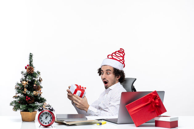年轻的商人带着圣诞老人帽子的年轻商人坐在办公室里看着白色背景上的礼物卖家人坐着