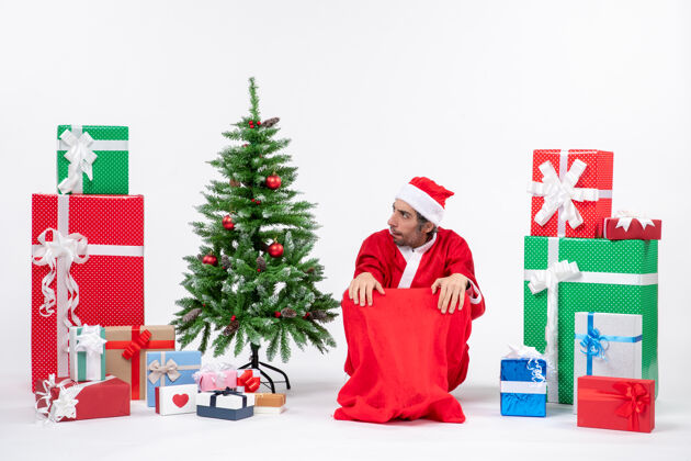 礼物悲伤的年轻人打扮成圣诞老人与礼物和装饰圣诞树上的白色背景圣诞树人年轻人