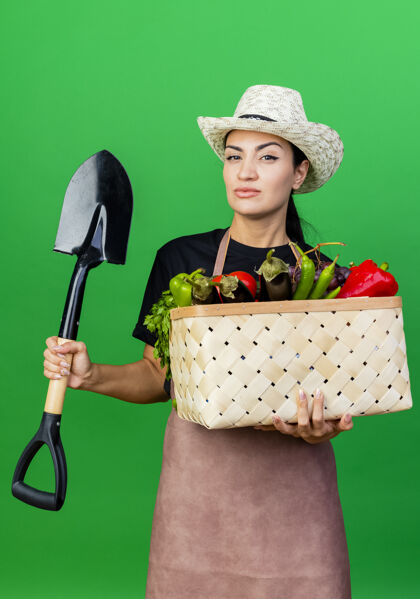 严肃的年轻漂亮的女园丁围着围裙 戴着帽子 手里拿着装满蔬菜的篮子和铲子 表情严肃绿色蔬菜围裙