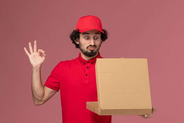 红色正面图穿着红色衬衫和披风的男信使拿着送食物的盒子打开它在浅粉色的墙上送货视图人