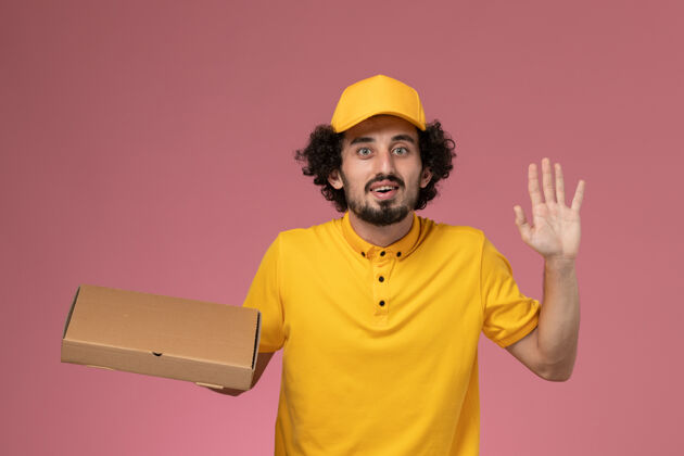 工作正面图：身穿黄色制服的男快递员拿着食品快递箱站在浅粉色的墙上工人前面制服