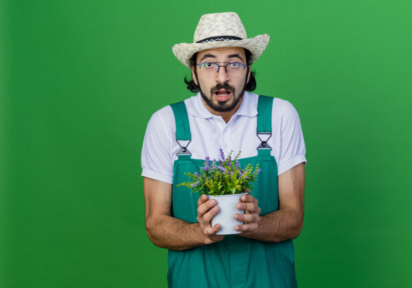 帽子年轻的留着胡子的园丁穿着连体衣戴着帽子拿着盆栽植物感到惊讶连身衣站着胡须