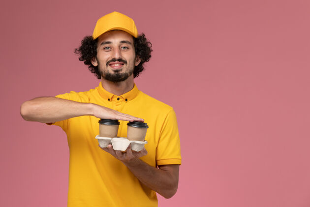 男正面图：身穿黄色制服和披风的男性信使手持棕色咖啡杯 微笑着站在粉红色的墙上人微笑咖啡