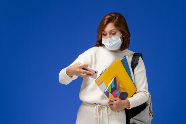 穿着正面图穿着白色球衣的年轻女学生戴着带包的面具和抄写本 在蓝色的墙上用手机人女人大学