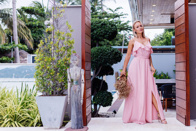 热带浪漫写真的粉红色晚装可爱的女人手持野花在豪华热带别墅外美丽的女性与花束女士户外人