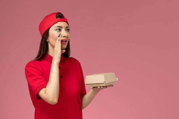 汉堡正面图：身穿红色制服和斗篷的年轻女快递员 手上拿着小包裹 在粉红色的墙上低语着视图快餐手
