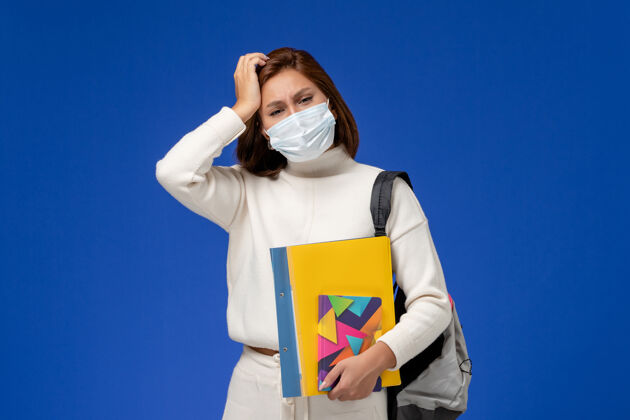 大学正面图身穿白色运动衫的年轻女学生戴着带包的面具 在蓝色的墙上写着头痛的字帖拥有漂亮面具
