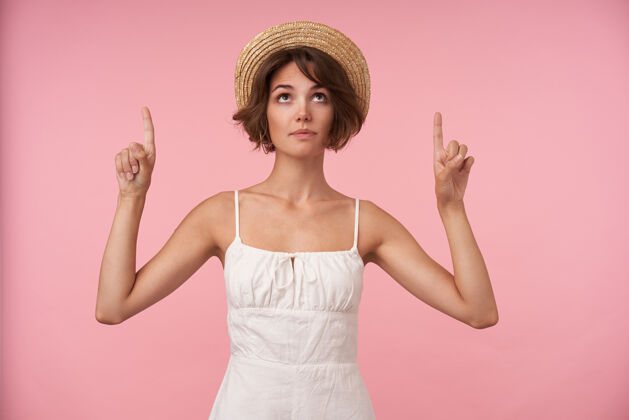棕色可爱的年轻女士 棕色短发 穿着白色优雅连衣裙 头戴草帽 摆姿势 向上看 食指抬起稻草女士手指