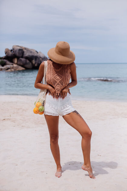 沙滩年轻漂亮的白种人 皮肤黝黑 穿着针织服装 戴着草帽 手拿一个装满水果 苹果 橘子 葡萄的线袋 在热带海滩上服装帽子天然