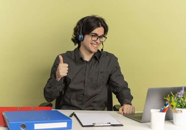 坐戴着耳机 戴着眼镜 面带微笑的年轻上班族坐在办公桌旁 手里拿着办公工具 竖起大拇指看着笔记本电脑工具空间工人