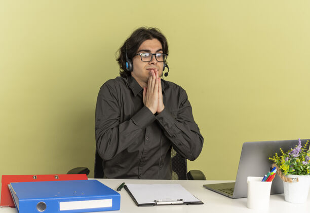 空间戴着耳机 戴着眼镜的年轻上班族坐在办公桌旁 手里拿着办公工具 看着笔记本电脑手坐耳机